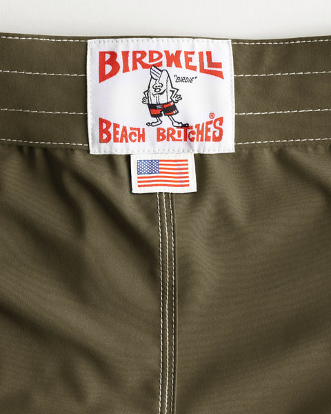 Close-up of Dark Olive 806 SurfStretch Side Pocket Boardshorts featuring a Birdwell Beach Britches label with a cartoon bird and a small American flag tag beneath it. White stitching stands out on the waistband and seam, showcasing these stretch boardshorts made in the USA.