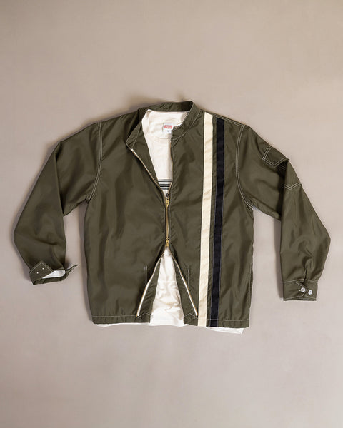 The Racing Jacket 26 - Dark Olive/Bone/Black, featuring vintage-inspired white and black stripes on the left side, is laid flat over a white T-shirt on a beige surface.