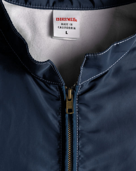 Close-up of the Racing Jacket 26 - Navy/Bone/Red, featuring a navy blue exterior with a visible zipper and contrasting white stitching. The interior label reads BIRDWELL MADE IN CALIFORNIA L in red and black text, emphasizing its American-made craftsmanship.