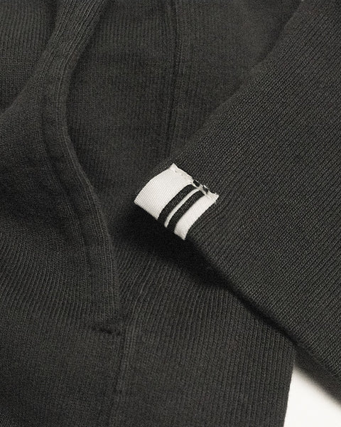 A close-up of the Harmony Hoodie in Faded Black, featuring a white tag with three black stripes sewn onto the sleeve cuff, lying flat on a white surface.
