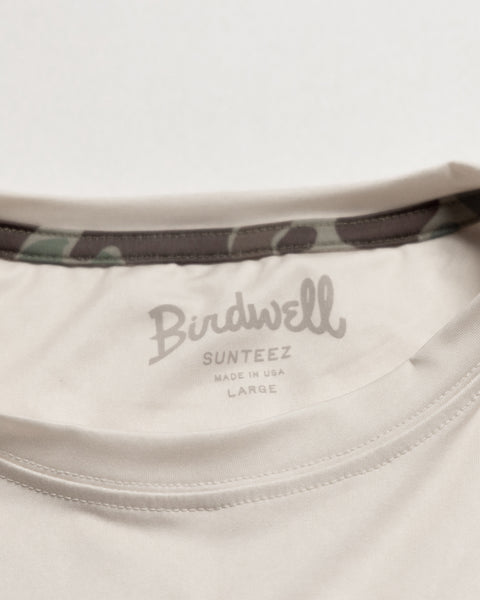 Close-up of the inside neck of a beige Sunteez Long Sleeve - Bone t-shirt, with BIRDWELL SUNTEEZ MADE IN USA LARGE printed on the inner collar and a hint of camouflage fabric lining. Crafted from moisture-wicking, performance stretch fabric for all-day comfort.