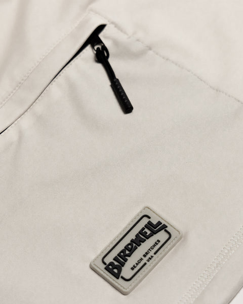 Close-up of the Sunteez Long Sleeve in Bone, showcasing beige performance stretch fabric with moisture-wicking properties for all-day comfort, and featuring a small zippered pocket above a rectangular Birdwell Beach Britches patch label stitched below the zipper.
