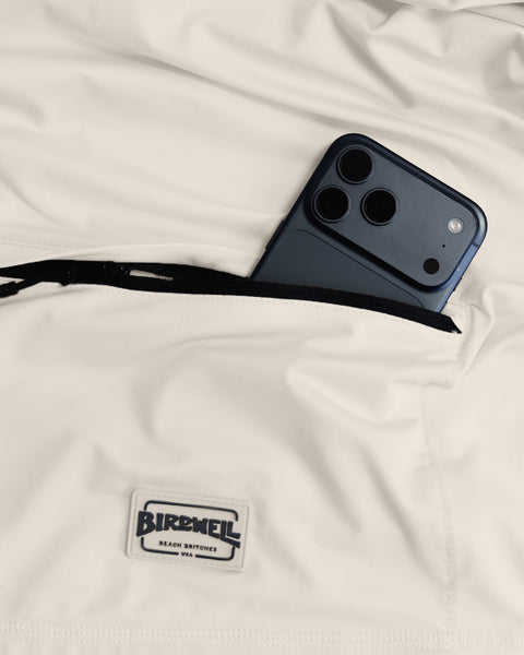 A black smartphone partially tucked into the zippered pocket of the cream-colored Sunteez Short Sleeve - Bone shirt, with a Birdwell Beach Britches label below the pocket.