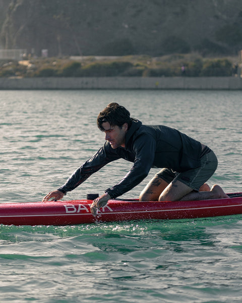 A person crouches on a red paddleboard, balancing over calm water near a rocky shoreline with greenery and a concrete wall in the background, wearing the Sunteez Hoodie - Black for UPF50+ sun protection.