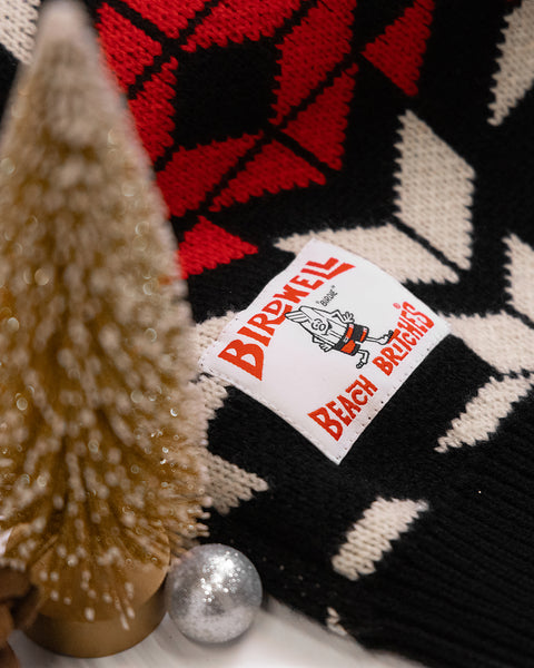 A close-up of the North Pole Sweater, featuring a red, black, and white geometric pattern. In the foreground are a gold bottlebrush tree and a silver ornament.