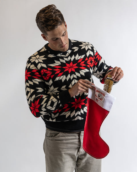 A man wearing a North Pole Sweater in black, white, and red puts a gold-wrapped gift and a chocolate bar into a red and white Christmas stocking against a plain white background.