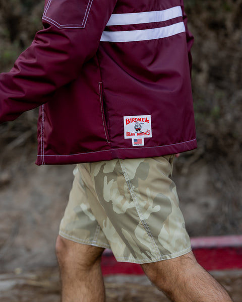 A person is walking outdoors near greenery and soil wearing the Comp Jacket 25 - Burgundy/White and beige camouflage shorts.