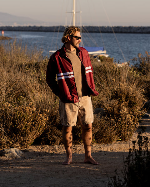 A man in sunglasses, barefoot on a sandy path near bushes with water and a docked sailboat behind him, wears the Comp Jacket 25 - Burgundy/White over a brown sweater and beige shorts.