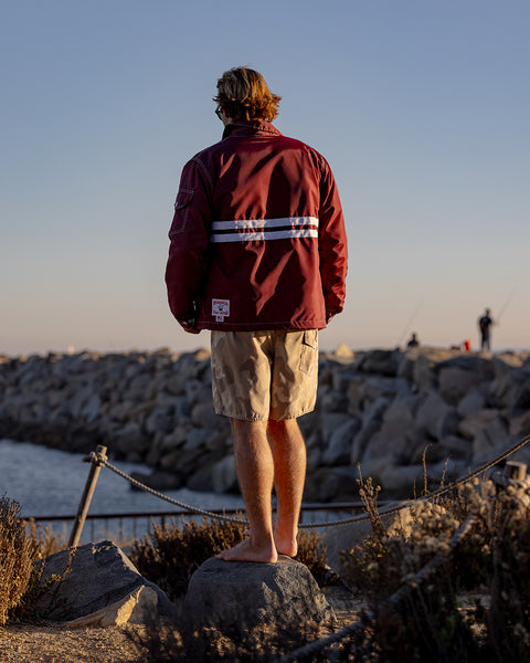 A person with short hair stands barefoot on rocks, facing away from the camera, wearing a Comp Jacket 25 - Burgundy/White and beige shorts. Behind them is a stone jetty and calm water under a clear sky.