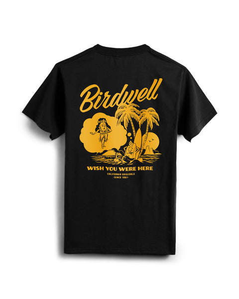 The Daydreamin' Birdie T-Shirt - Black is a relaxed fit, heavyweight cotton tee with yellow beach scene graphics—featuring a woman, palm trees, surfboards, ocean—and the iconic Birdwell text: “Wish you were here. California originals since 1961.”.