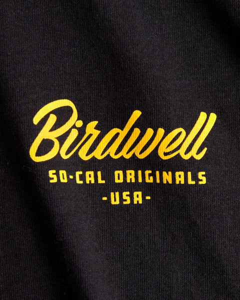 Close-up of the black fabric from the Daydreamin' Birdie T-Shirt, featuring yellow Birdwell SO-CAL ORIGINALS - USA - text. The design appears printed or embroidered on this heavyweight cotton tee.