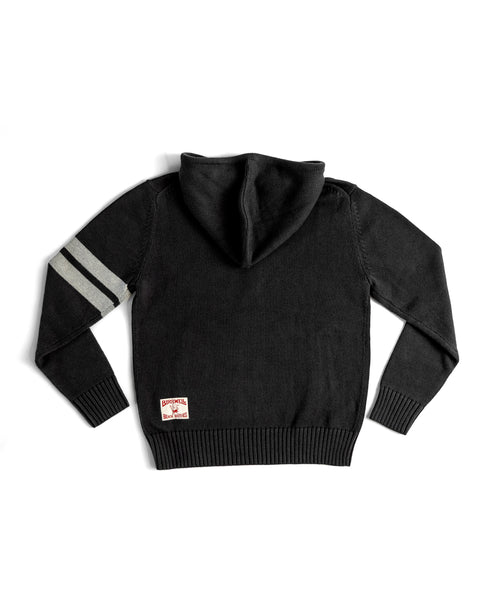 The Comp Zip Sweater - Black/Gray is shown laid flat, back facing up. It features two gray stripes on the left sleeve and a small red-text patch near the bottom hem. Made from 100% cotton, this midweight knit offers comfort.