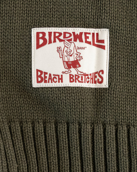 A close-up of the Birdwell Beach Britches label sewn onto the Comp Stripe Sweater - Dark Olive/Bone, made from 100% cotton knit, featuring a cartoon bird in swim trunks and bold red text on a white background.