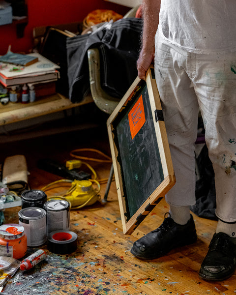 Wearing a paint-splattered BENT Comp Jacket - Black, a person holds framed artwork in a messy, paint-filled studio with art supplies, paint cans, and tools scattered across the wooden floor.