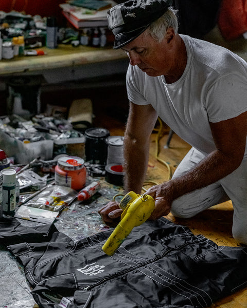 A man in a white t-shirt and cap kneels, using a yellow heat gun on a black BENT Comp Jacket. He’s surrounded by paint cans and art supplies in a cluttered, creative workspace.