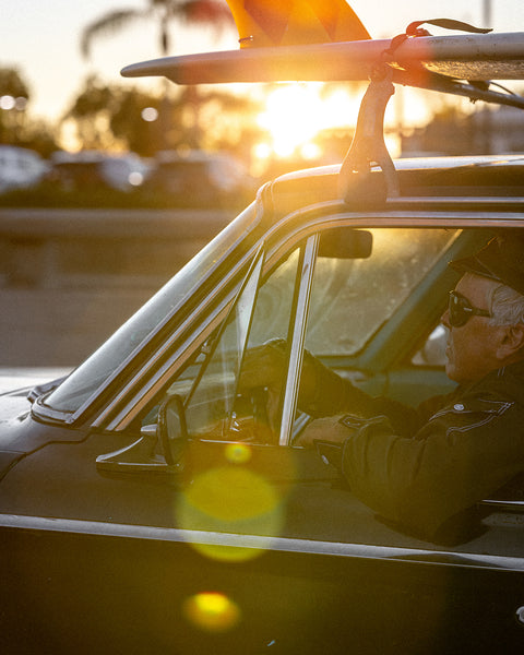 A man in sunglasses and a BENT Comp Jacket - Black drives a vintage car with a surfboard on the roof at sunset, sunlight and lens flares brightening the scene.