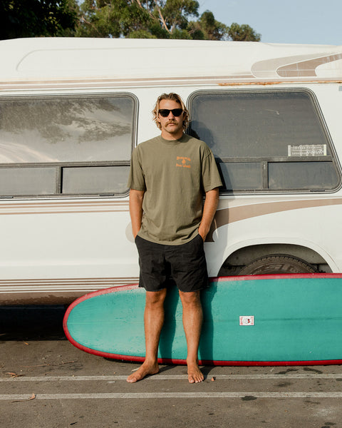 A barefoot man in sunglasses and a License Plate T-Shirt - Army/Orange stands in front of a white camper van. Black shorts, a green and red surfboard on the ground, and trees are visible in the background.