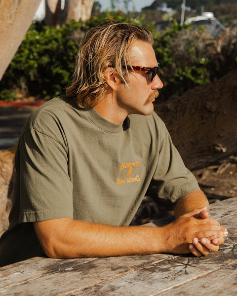 A man with blonde hair and a mustache, wearing sunglasses and a License Plate T-Shirt - Army/Orange, sits at a weathered wooden table outdoors with greenery and sunlight in the background.