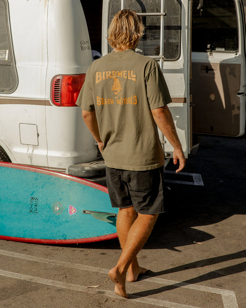 A person with blond hair, wearing a License Plate T-Shirt - Army/Orange and black shorts, stands barefoot near a white van and blue surfboard in a sunlit parking lot.
