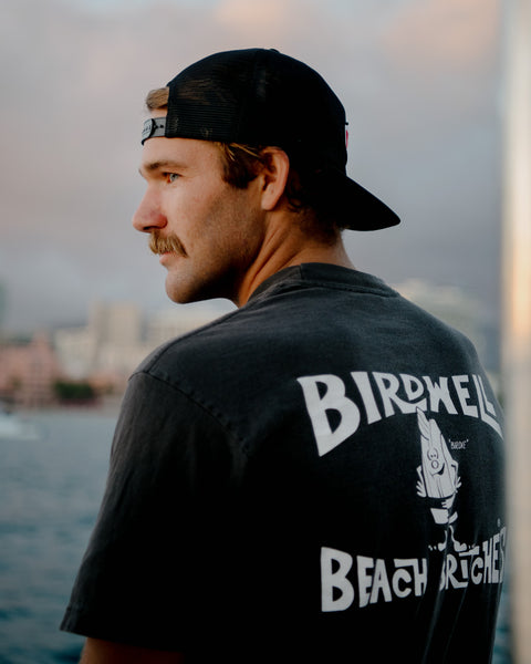 A man with a mustache, wearing a black cap backward and a Birdwell Beach Britches License Plate T-Shirt - Black/White, stands near water looking to the side, with a blurred cityscape in the background.