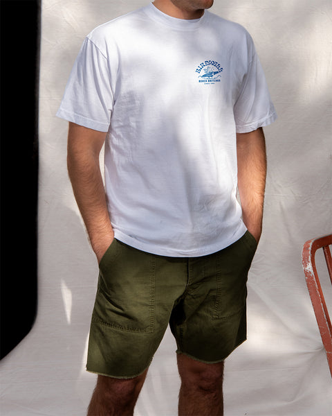 A person wears the Surfin' Birdie T-Shirt - White/Blue and olive green shorts, standing with hands in pockets against a neutral background. Only their torso and upper legs are visible; their face is not shown.