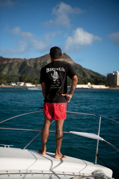 Wearing the 61 T-Shirt - White/Black and red shorts, a person stands on a boat with their back to the camera, facing the ocean. Green mountains, buildings, and a partly cloudy blue sky appear in the background.