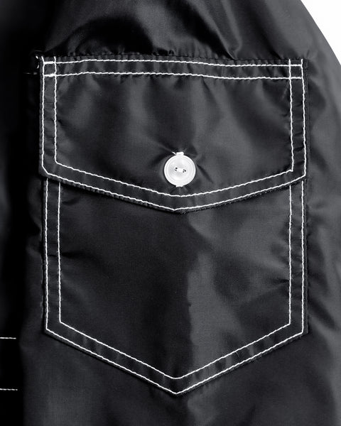 A close-up of a black fabric pocket on a BENT Comp Jacket - Black, featuring angular edges, visible white stitching, a pointed flap, and a centered white button.